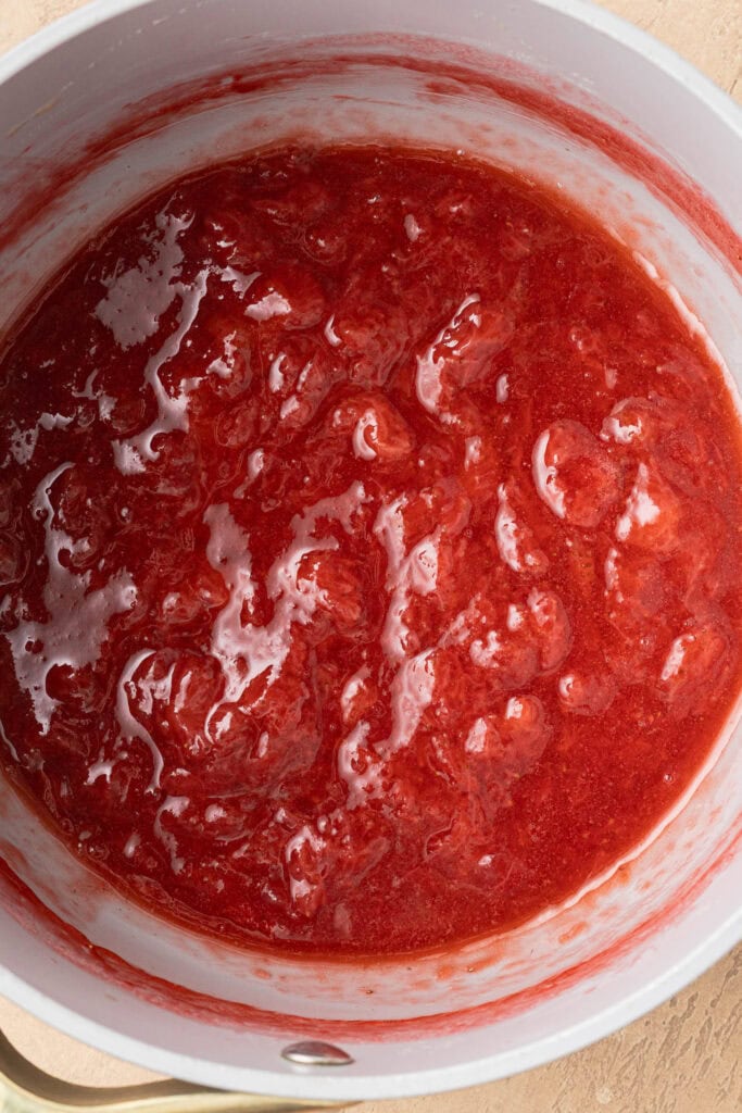 Bright red strawberry sauce with a thick, chunky texture in a white saucepan, viewed from above.