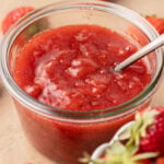 A glass jar of strawberry jam with a spoon inside, surrounded by fresh strawberries.