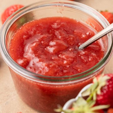 A glass jar of strawberry jam with a spoon inside, surrounded by fresh strawberries.