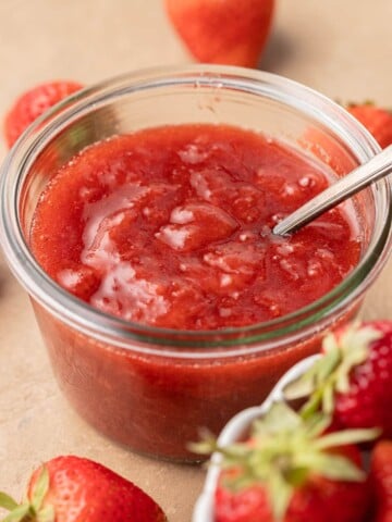 A glass jar of strawberry jam with a spoon inside, surrounded by fresh strawberries.