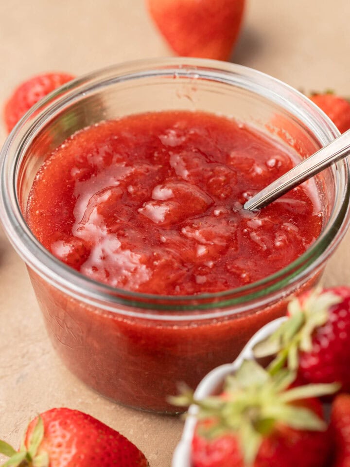 A glass jar of strawberry jam with a spoon inside, surrounded by fresh strawberries.