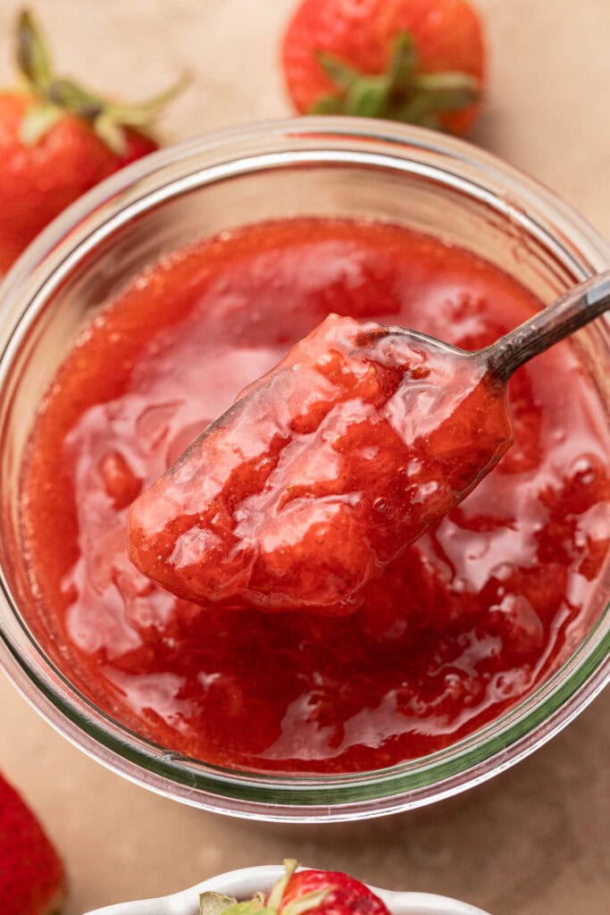 A spoonful of strawberry jam held over a glass jar, with fresh strawberries in the background.
