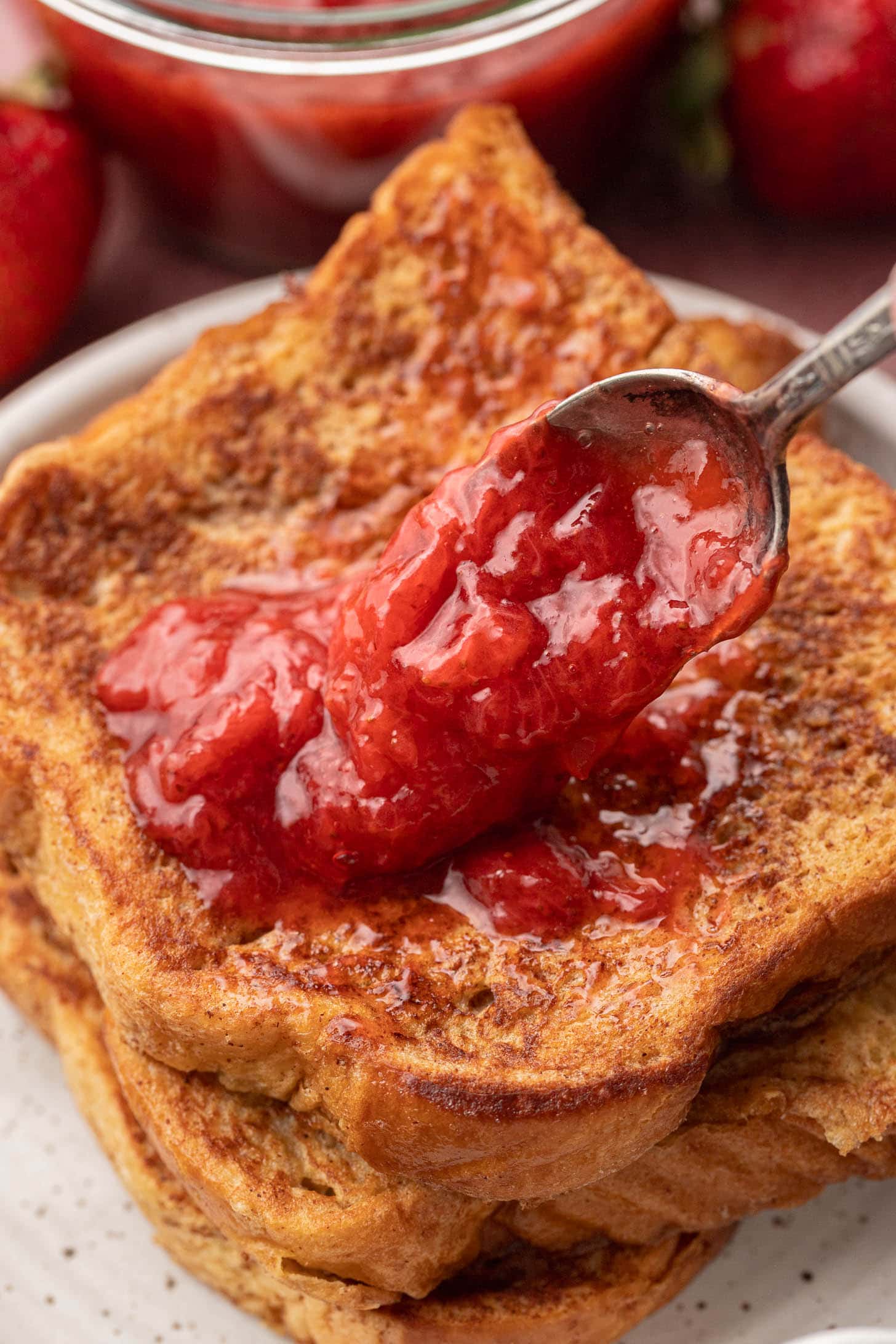 A spoon spreads strawberry jam over a stack of golden brown French toast slices on a plate.