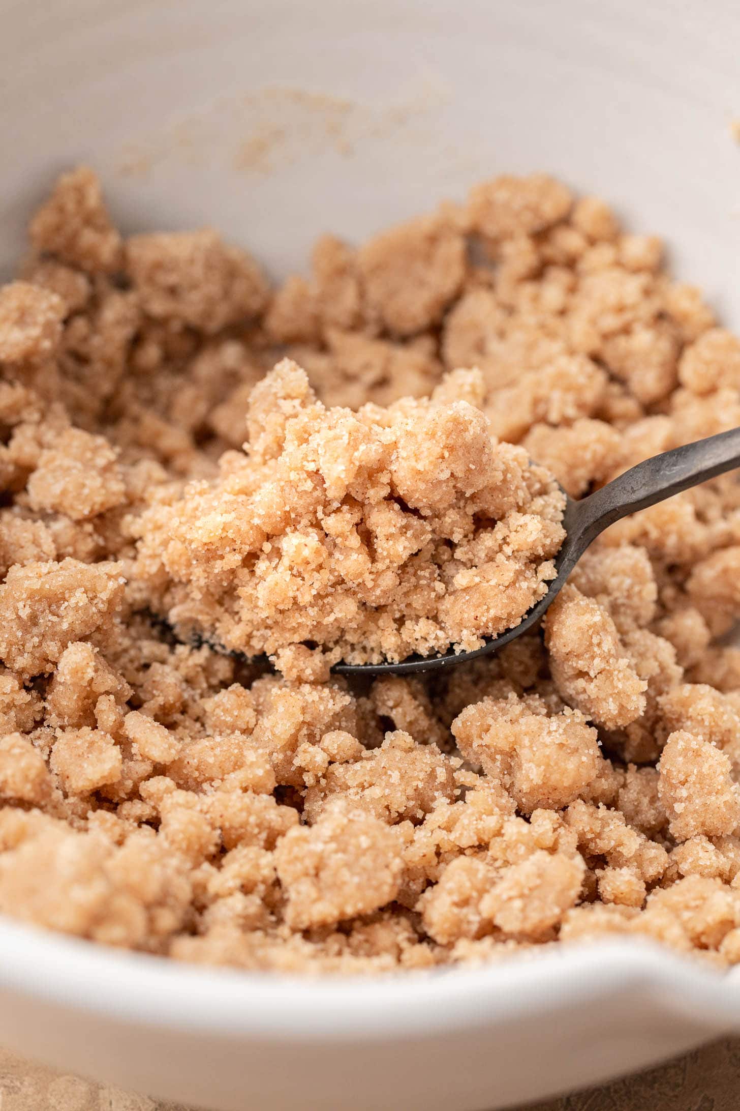 A spoon scoops crumbly brown sugar and flour mixture from a white bowl.