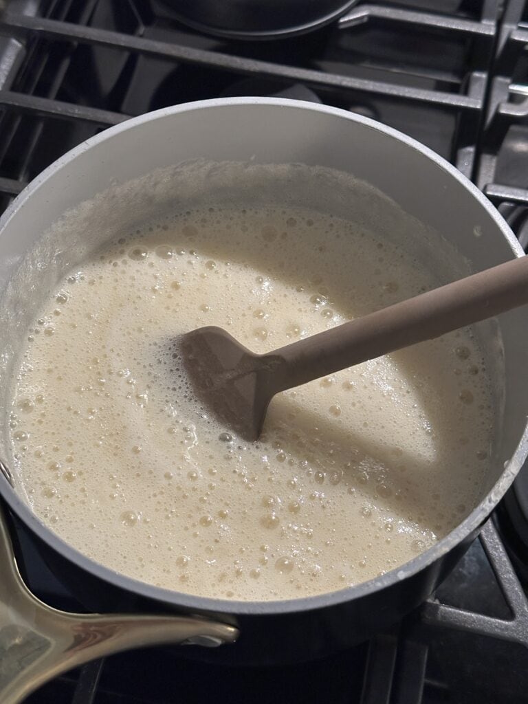 A brown spatula stirring a frothy, bubbling mixture in a saucepan on a stovetop.