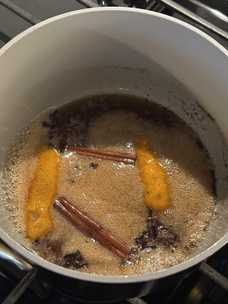 A pot on a stove with simmering liquid, orange peels, cinnamon sticks, and star anise floating.