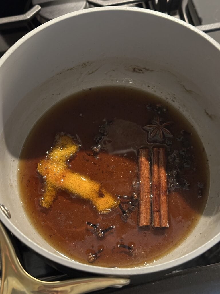 A pot on a stove with spiced syrup, orange peel, cinnamon sticks, cloves, and a star anise.