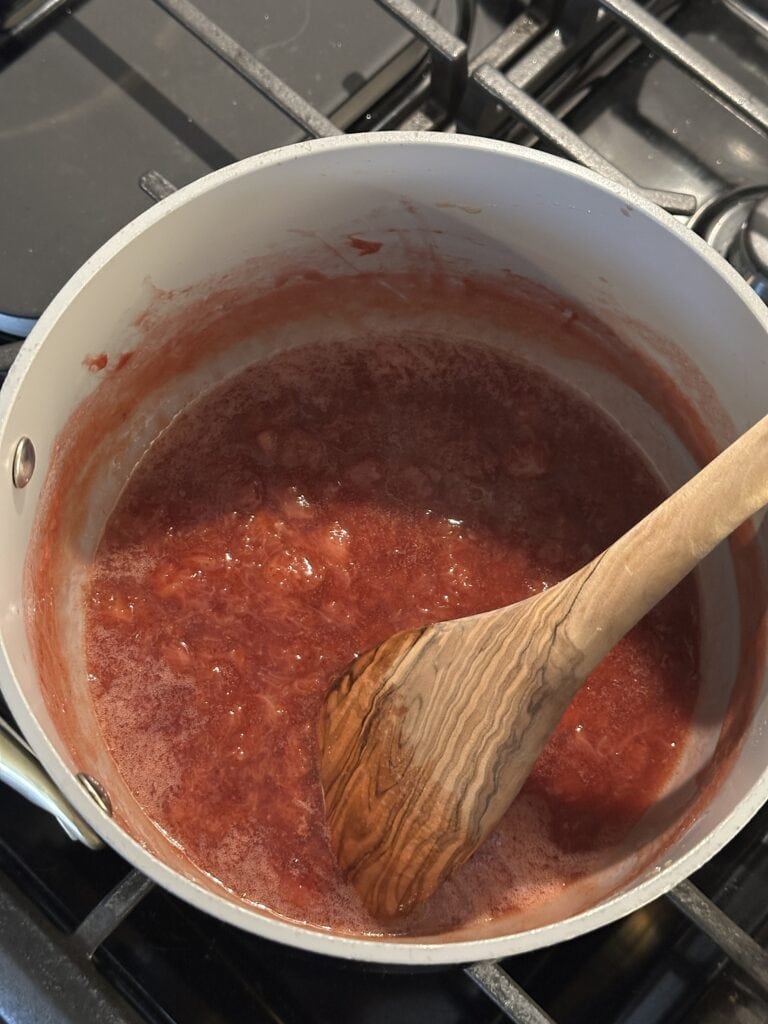 A wooden spoon stirring red fruit jam in a white saucepan on a stovetop.