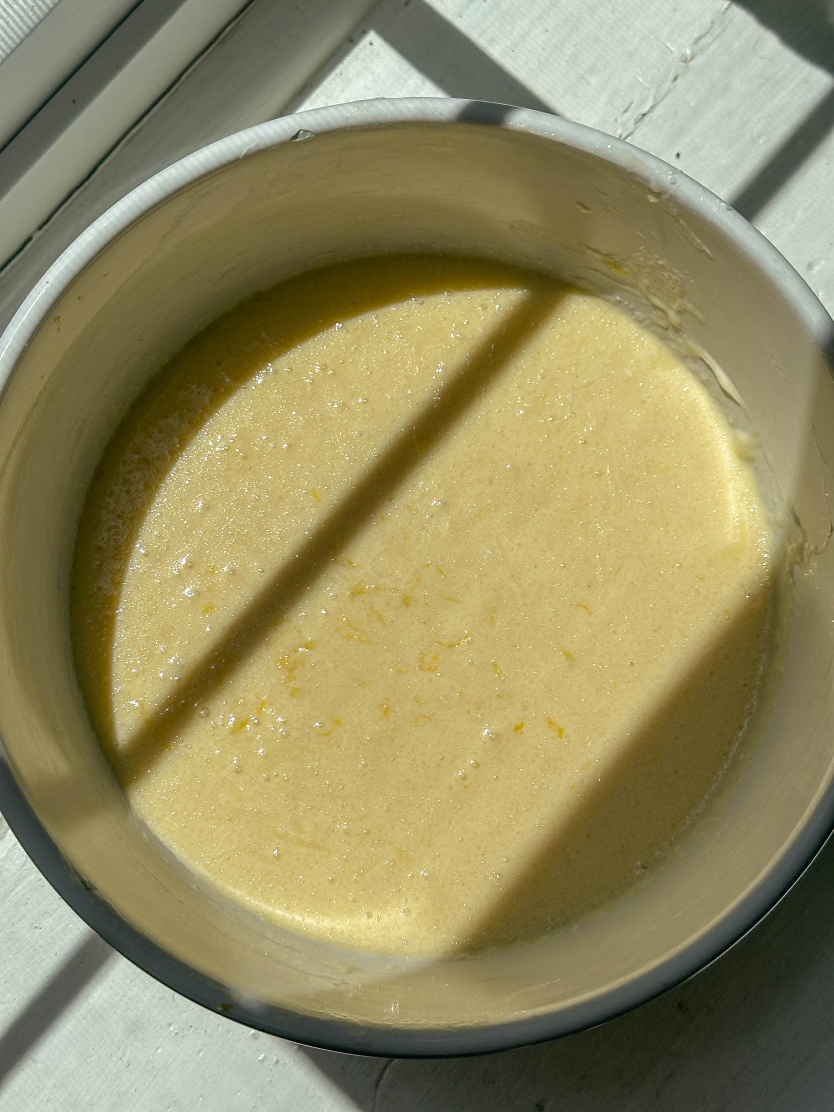 A mixing bowl with yellow muffin batter for lemon muffins sits in sunlight with window shadows across it.