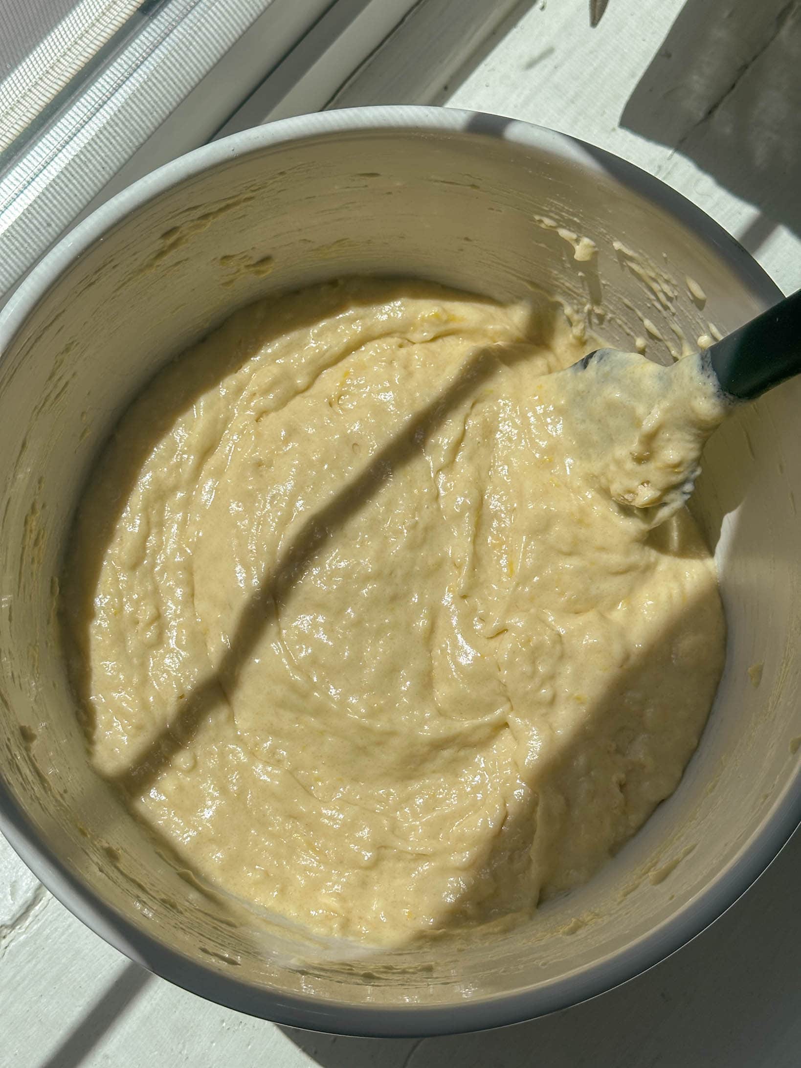 A mixing bowl of pale lemon muffin batter with a spatula, lit by sunlight streaming through window blinds.