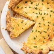 A sliced quiche with chives on top, in a white pie dish, next to a wedge of cheese and a bowl of chives.