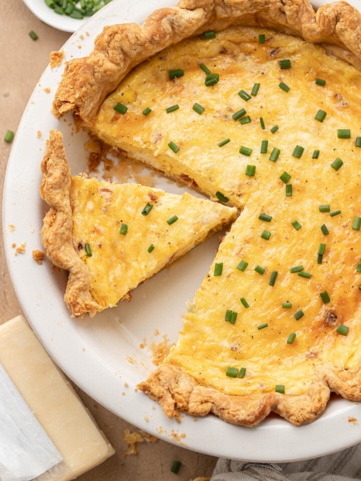 A sliced quiche with chives on top, in a white pie dish, next to a wedge of cheese and a bowl of chives.