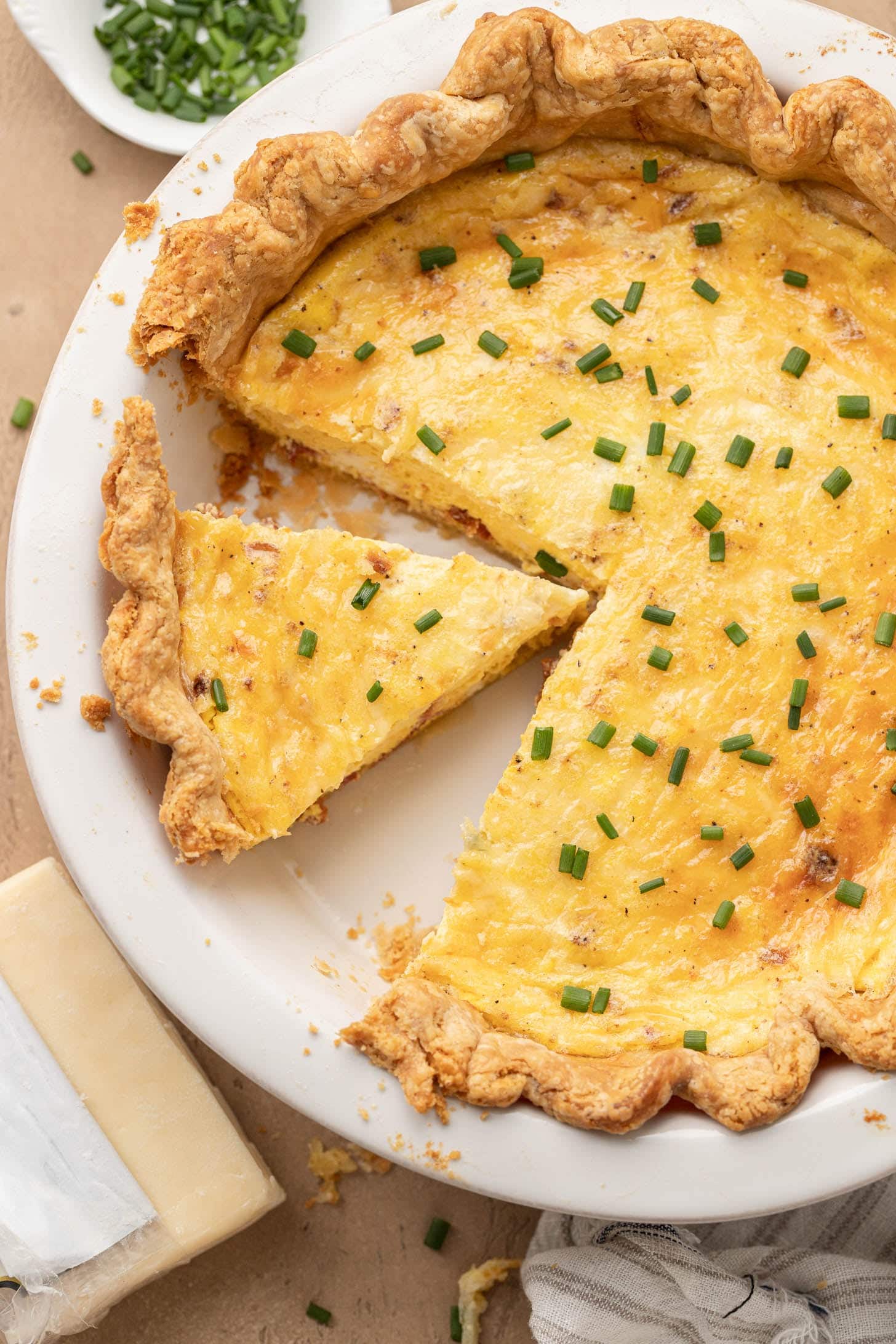 A sliced quiche with chives on top, in a white pie dish, next to a wedge of cheese and a bowl of chives.