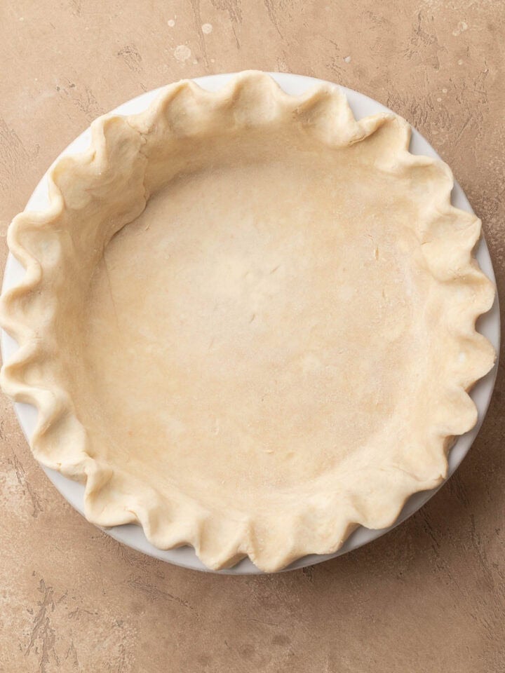 Unbaked pie crust with crimped edges in a white pie dish on a tan surface.