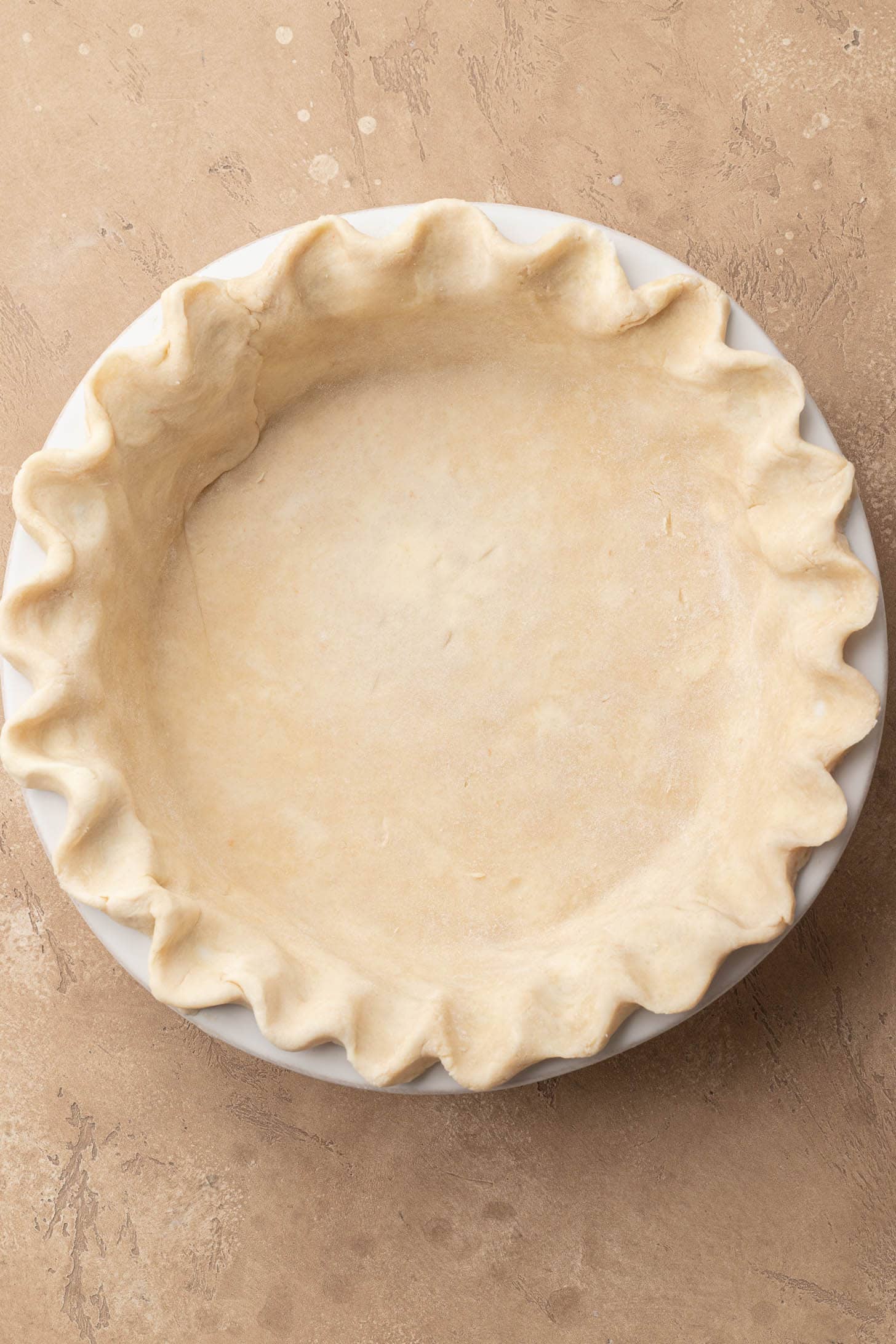 Unbaked pie crust with crimped edges in a white pie dish on a tan surface.