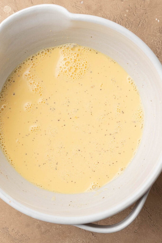 A white bowl filled with a pale yellow, whisked egg mixture with small bubbles and specks of pepper.