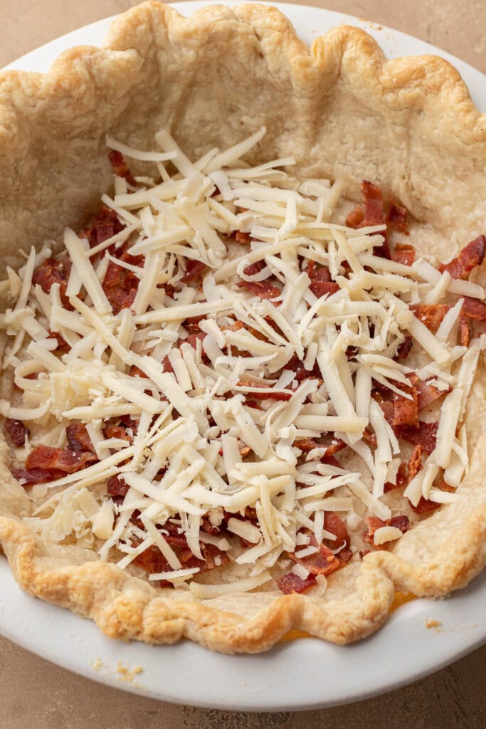 Unbaked pie crust filled with shredded cheese and chopped bacon in a white dish.