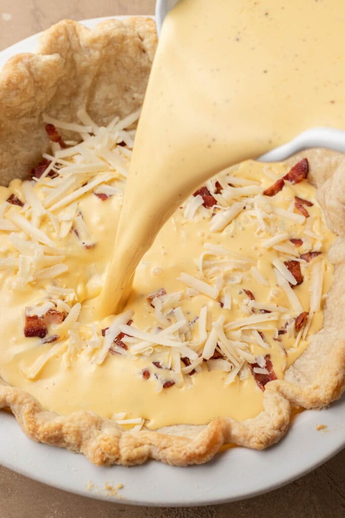 Pie crust filled with cheese and bacon is topped with creamy egg mixture being poured in.