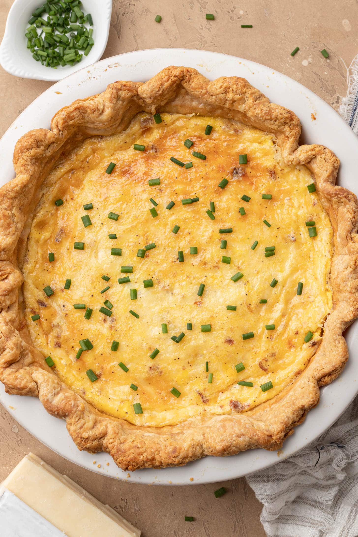 A baked quiche garnished with chopped chives on a plate, with extra chives in a small dish nearby.