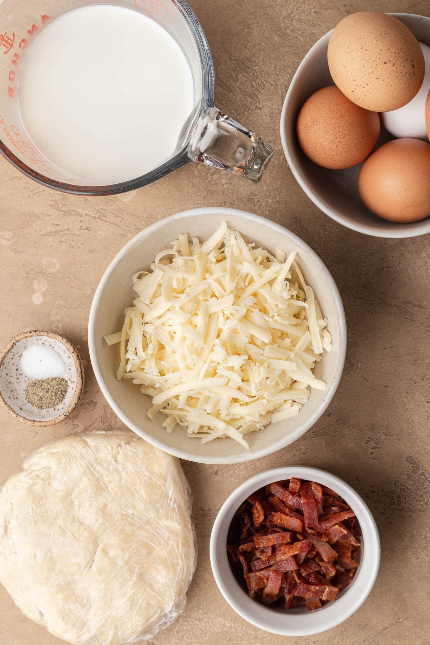 Overhead view of eggs, shredded cheese, cooked bacon, pie dough, milk, and salt and pepper on a tan surface.