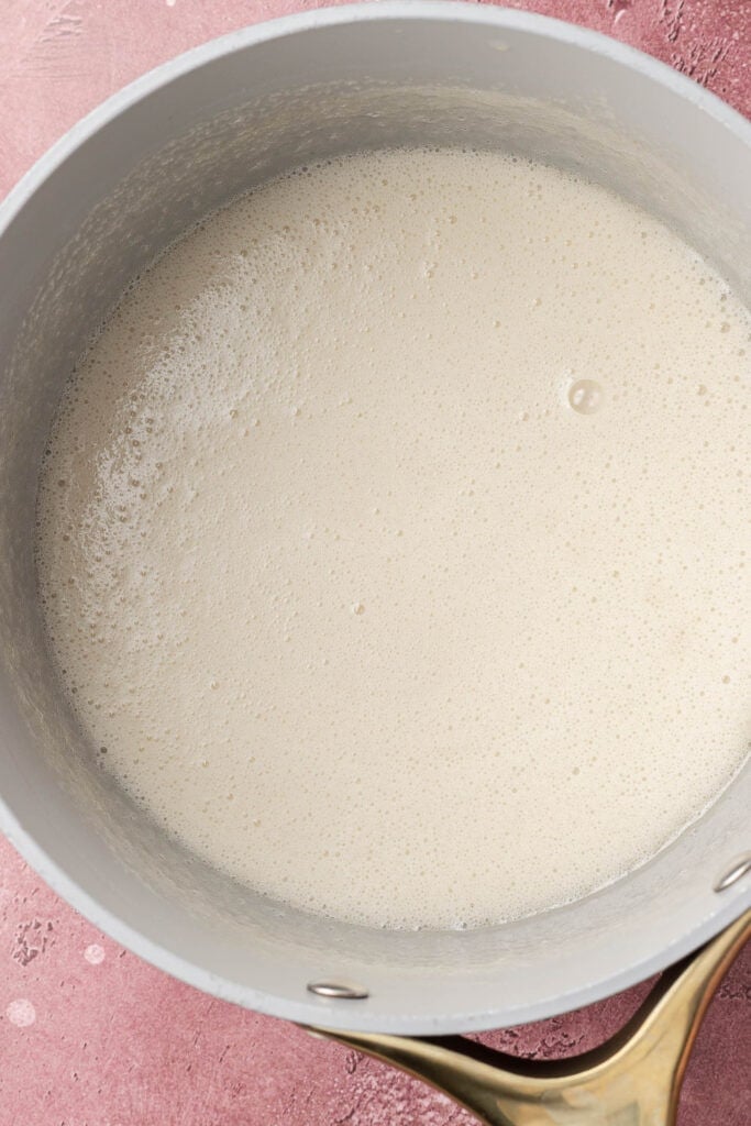 A saucepan filled with a creamy, frothy white liquid on a pink textured surface.