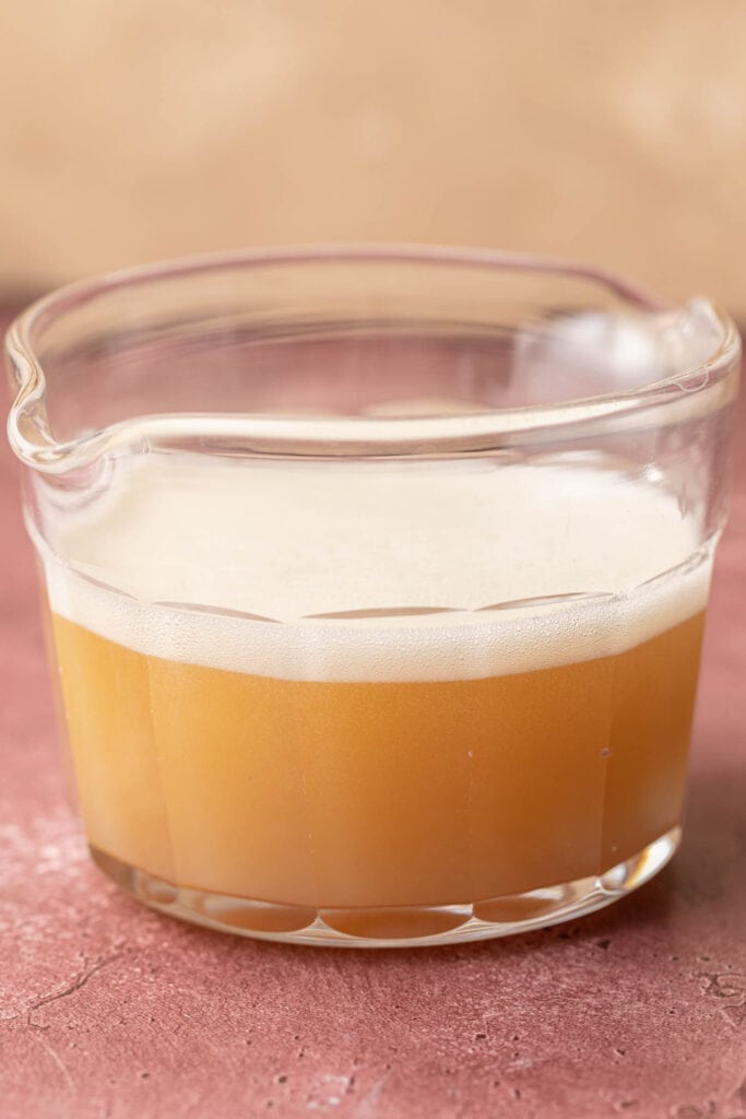 A glass measuring cup filled with a frothy, light golden liquid on a textured pink surface.