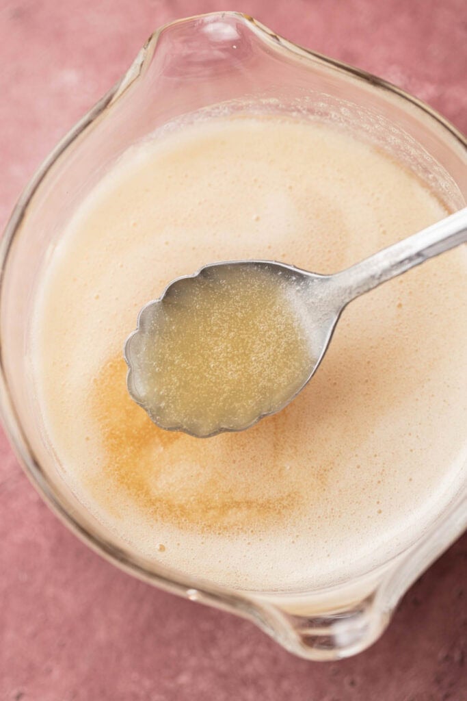 A spoon holding foamy buttermilk mixture over a glass bowl on a pink surface.