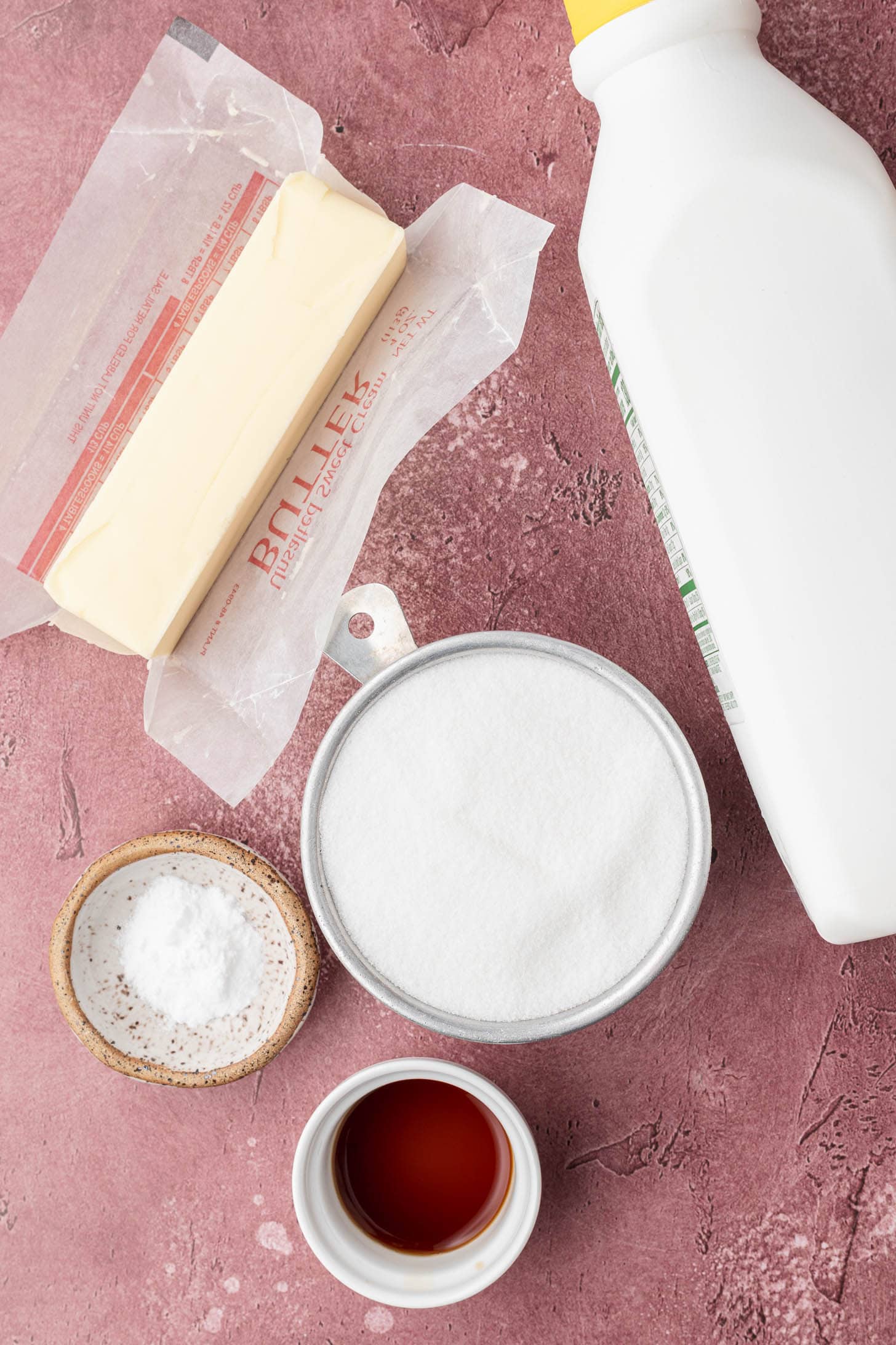 Stick of butter, bottle of buttermilk, bowl of sugar, small bowl of baking soda, and cup of vanilla extract on pink surface.