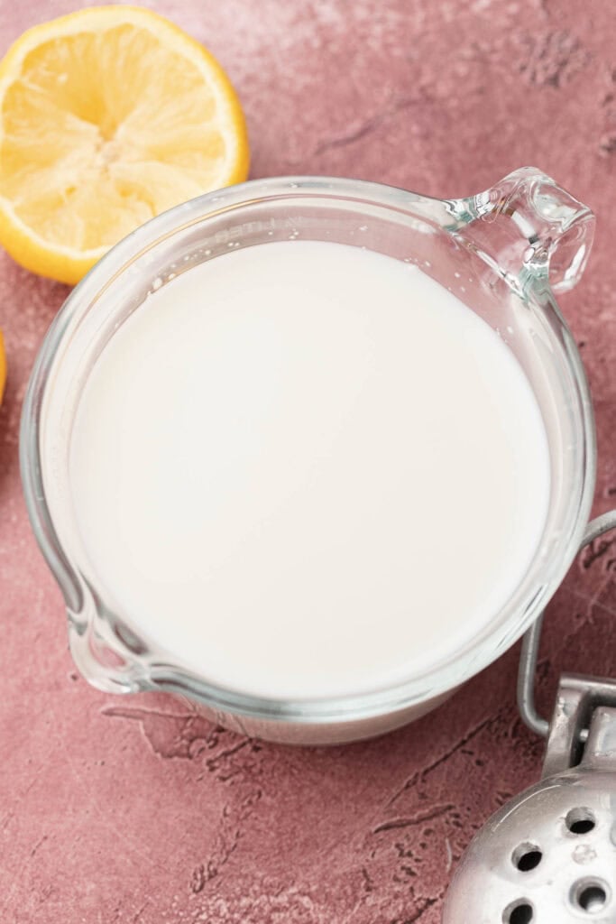 A glass measuring cup of buttermilk next to lemon halves on a pink textured surface.