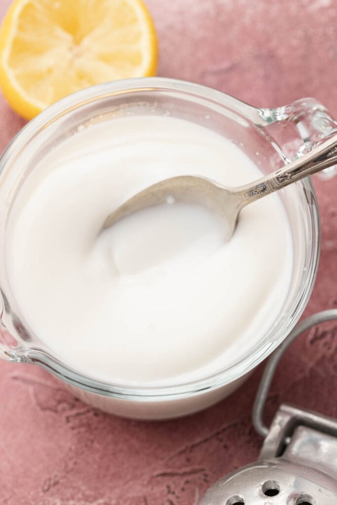 A spoon in a glass measuring cup of creamy buttermilk, with a lemon half and juicer nearby on a pink surface.