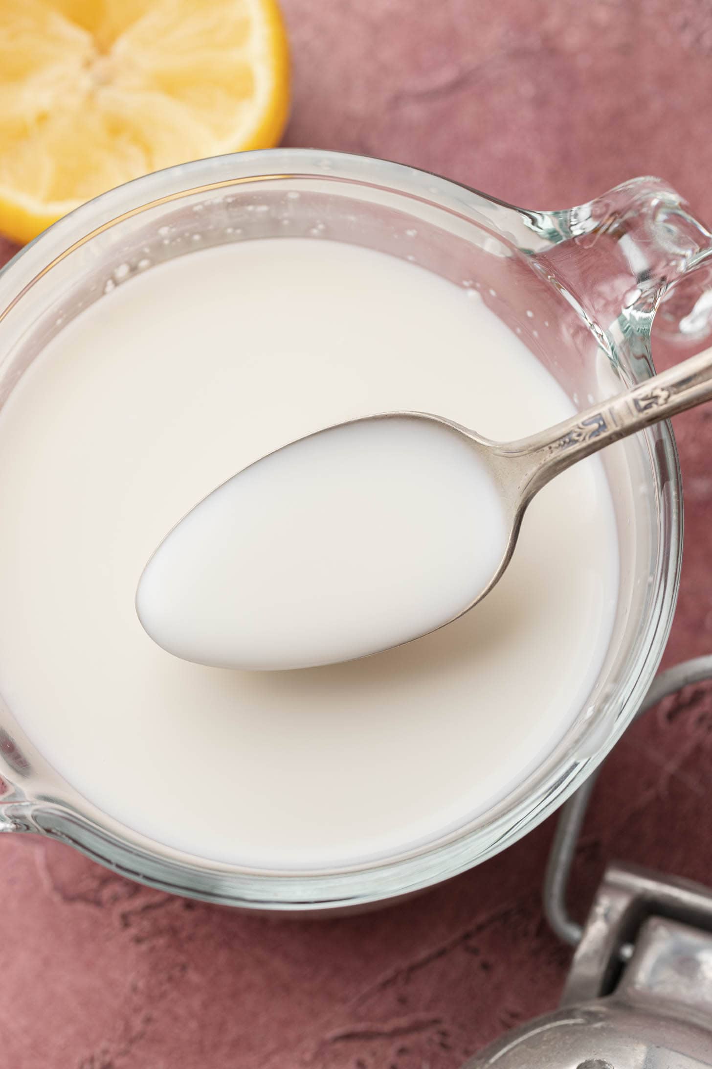 A spoon rests in a glass measuring cup filled with a white liquid, with a lemon wedge nearby.