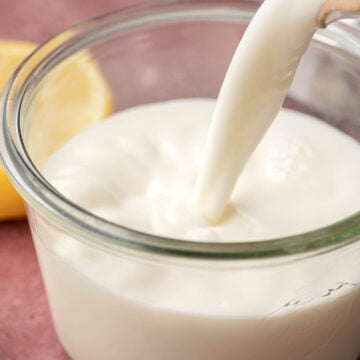 buttermilk being poured into a clear glass jar, with a lemon in the background on a pink surface.