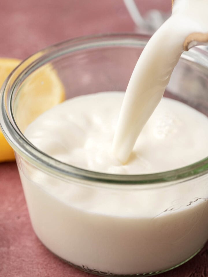 buttermilk being poured into a clear glass jar, with a lemon in the background on a pink surface.