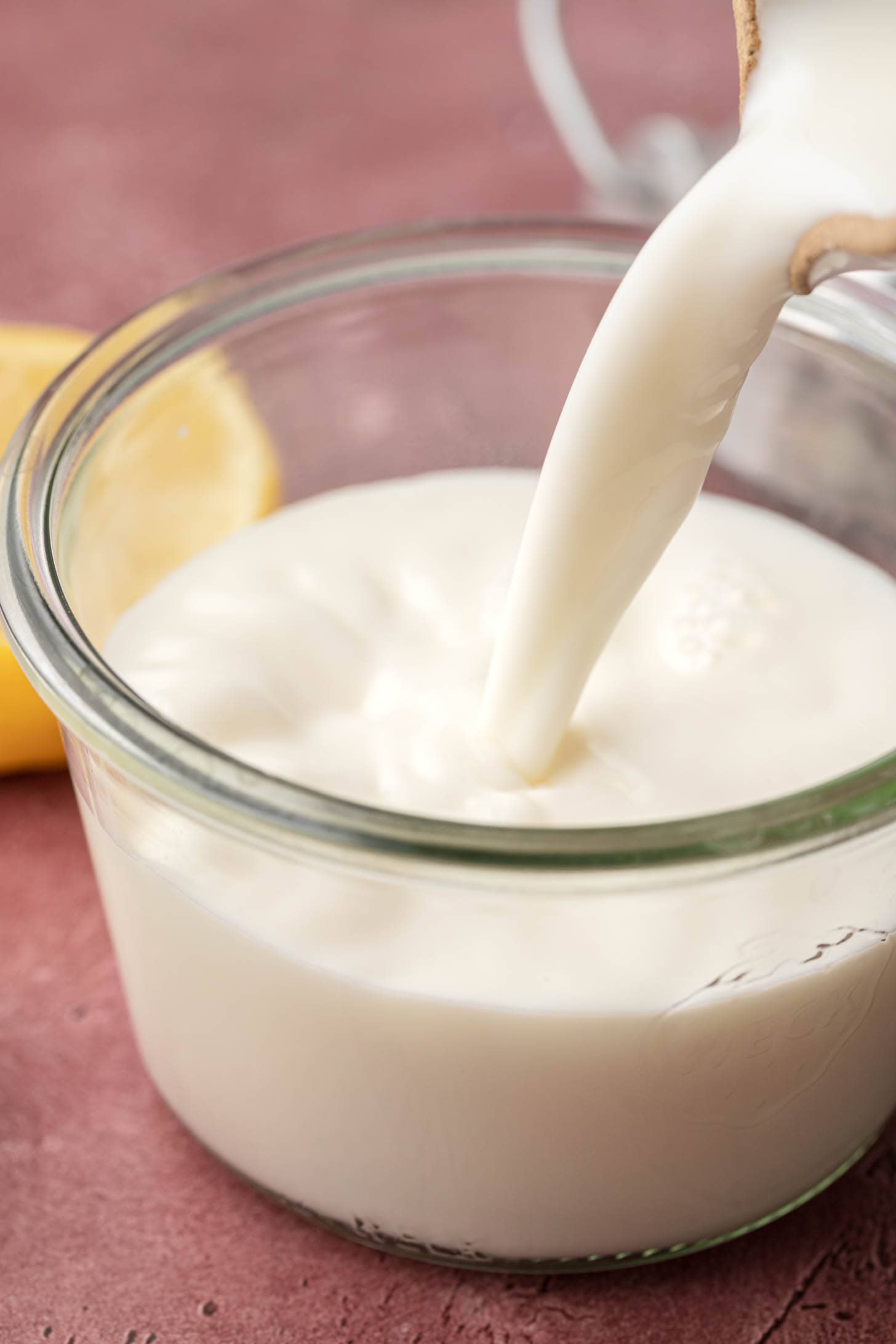buttermilk being poured into a clear glass jar, with a lemon in the background on a pink surface.