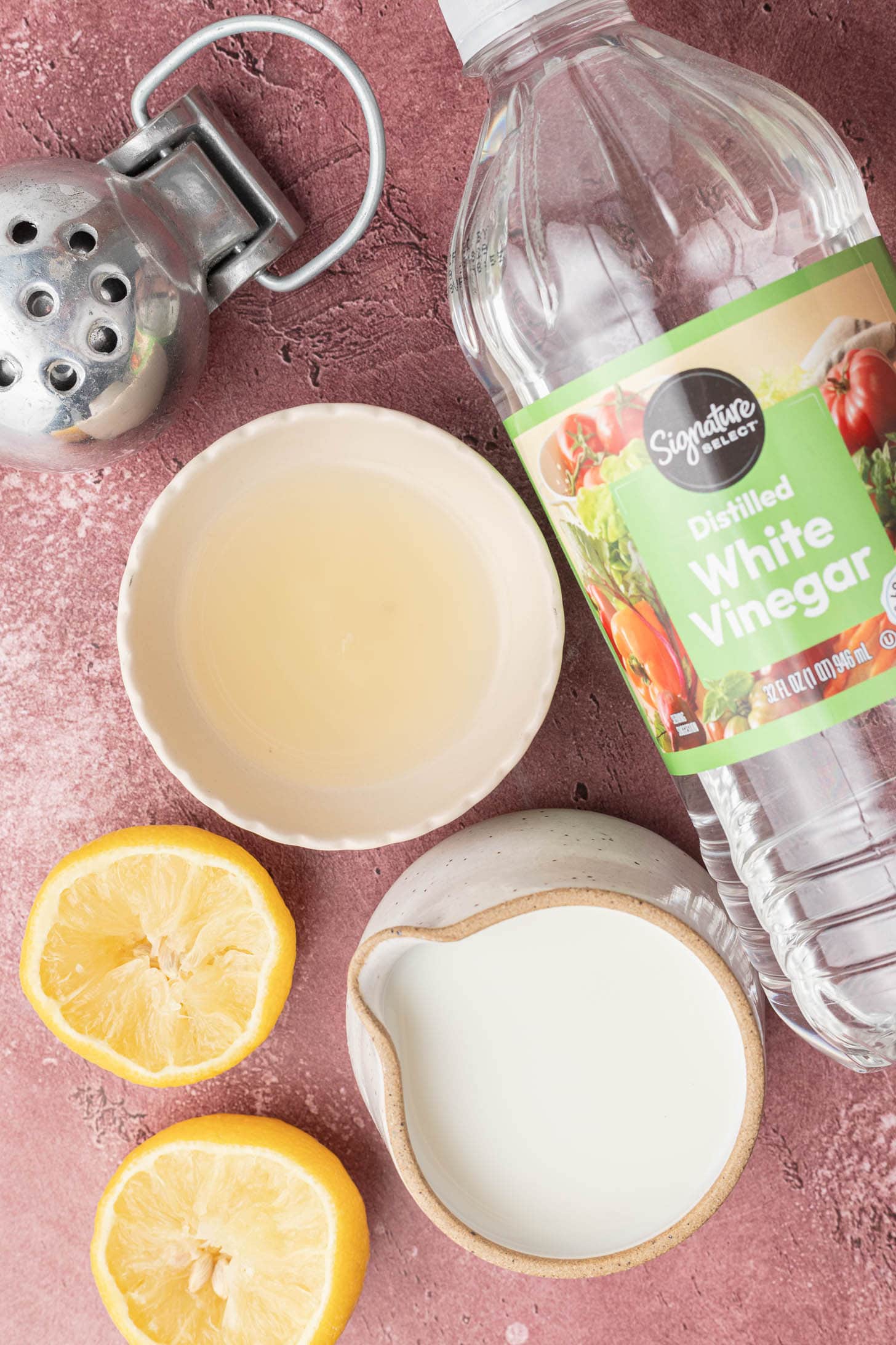 White vinegar, sliced lemon, a small bowl of lemon juice, a pitcher of milk, and a metal juicer on a pink surface.