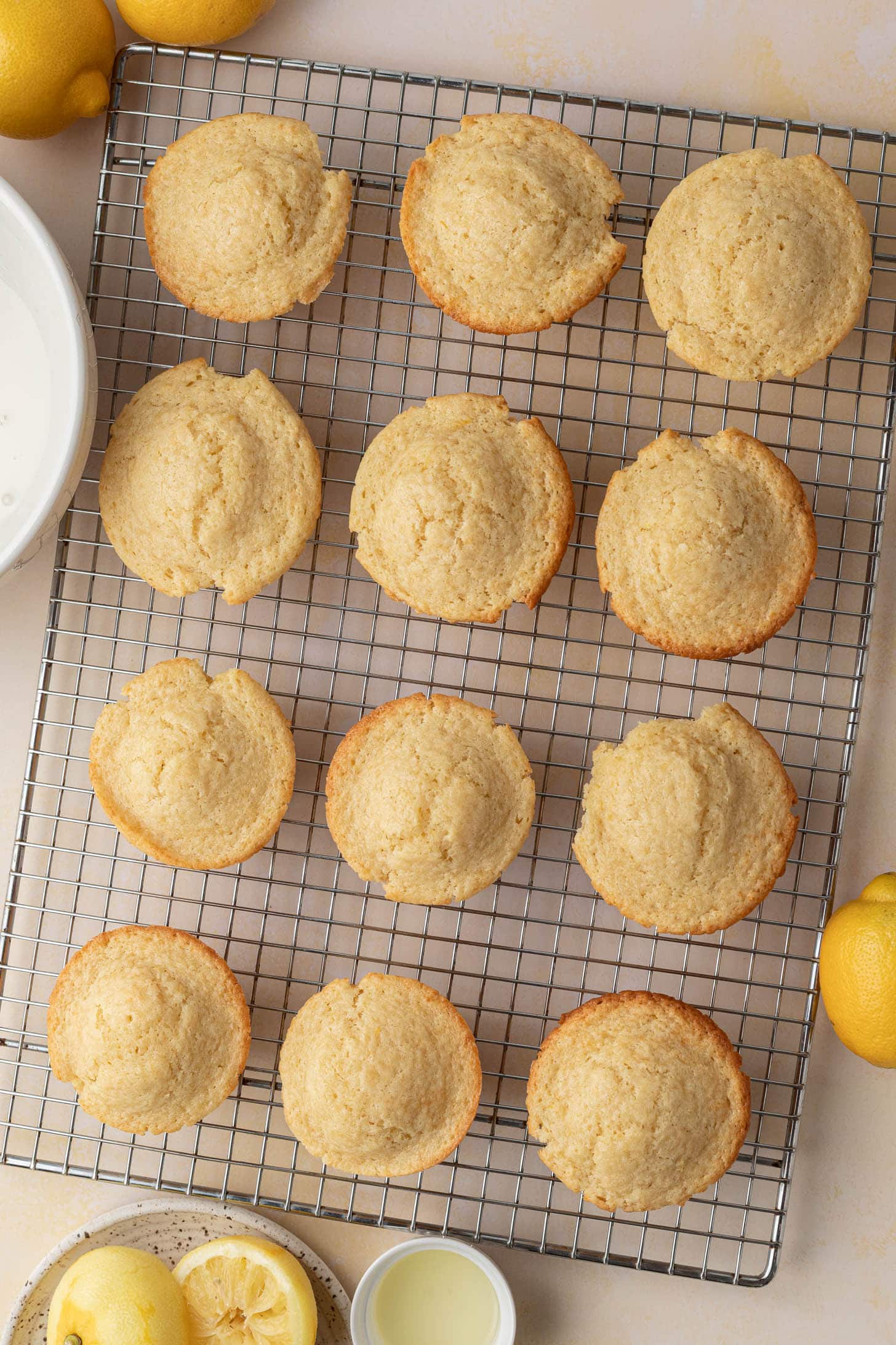 Twelve baked lemon muffins cooling on a wire rack, with lemons and bowls nearby.