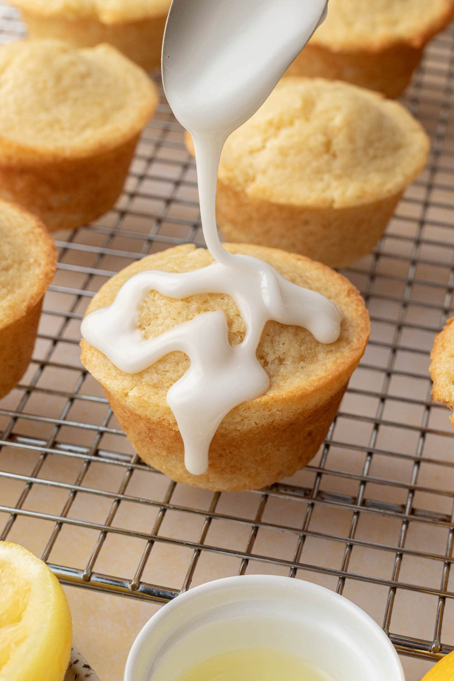 A spoon drizzles white icing over a muffin on a cooling rack, with more muffins in the background.