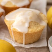 Lemon muffin topped with white glaze, surrounded by fresh lemons on a wooden surface.