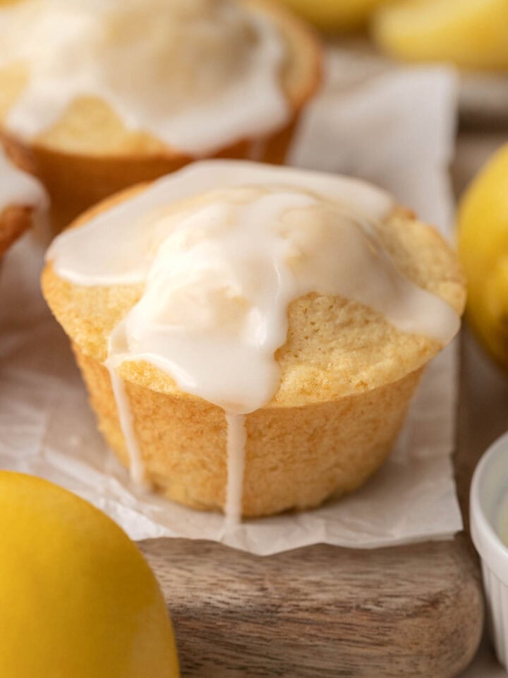 Lemon muffin topped with white glaze, surrounded by fresh lemons on a wooden surface.