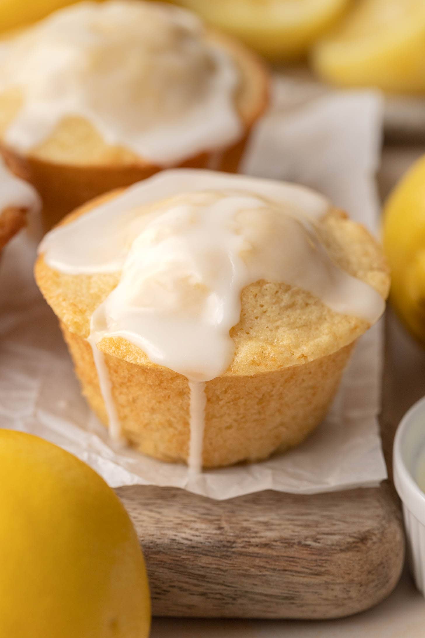 Lemon muffin topped with white glaze, surrounded by fresh lemons on a wooden surface.