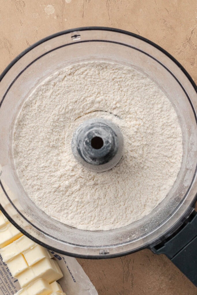 Top view of flour in a food processor bowl next to cubed butter on a tan surface.