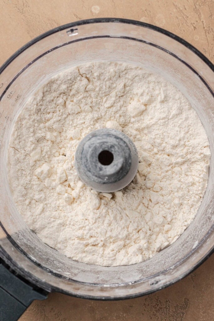 Flour and butter mixture in a food processor bowl, viewed from above, on a beige countertop.