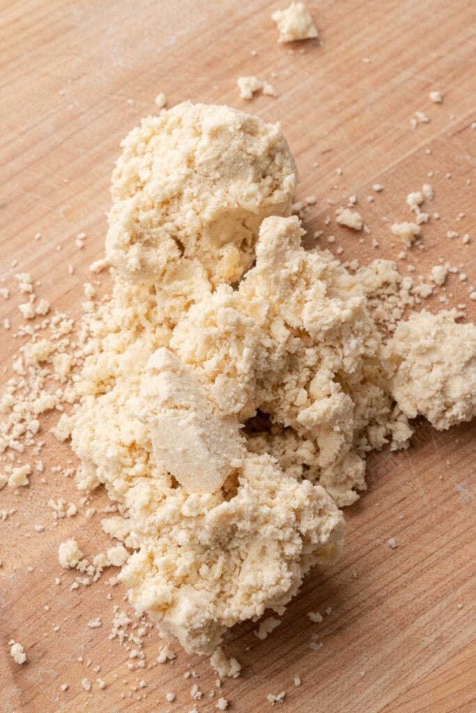 Coarse, crumbly dough mixture on a wooden surface, with small chunks and loose crumbs scattered around.