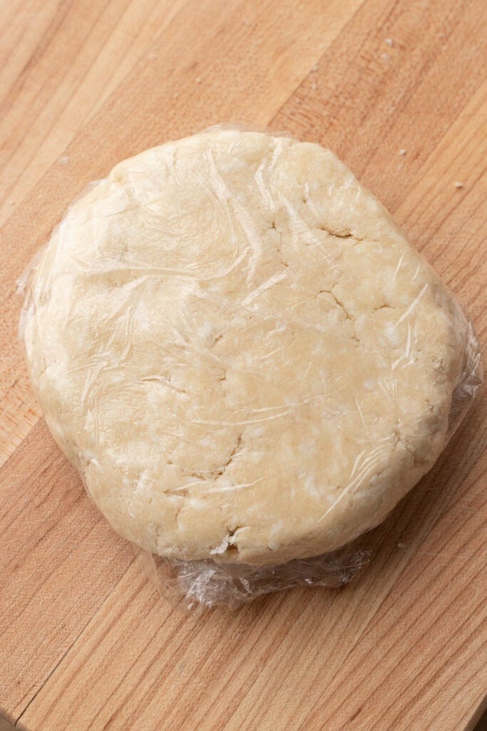 Round disk of pie dough wrapped in plastic wrap on a wooden surface.