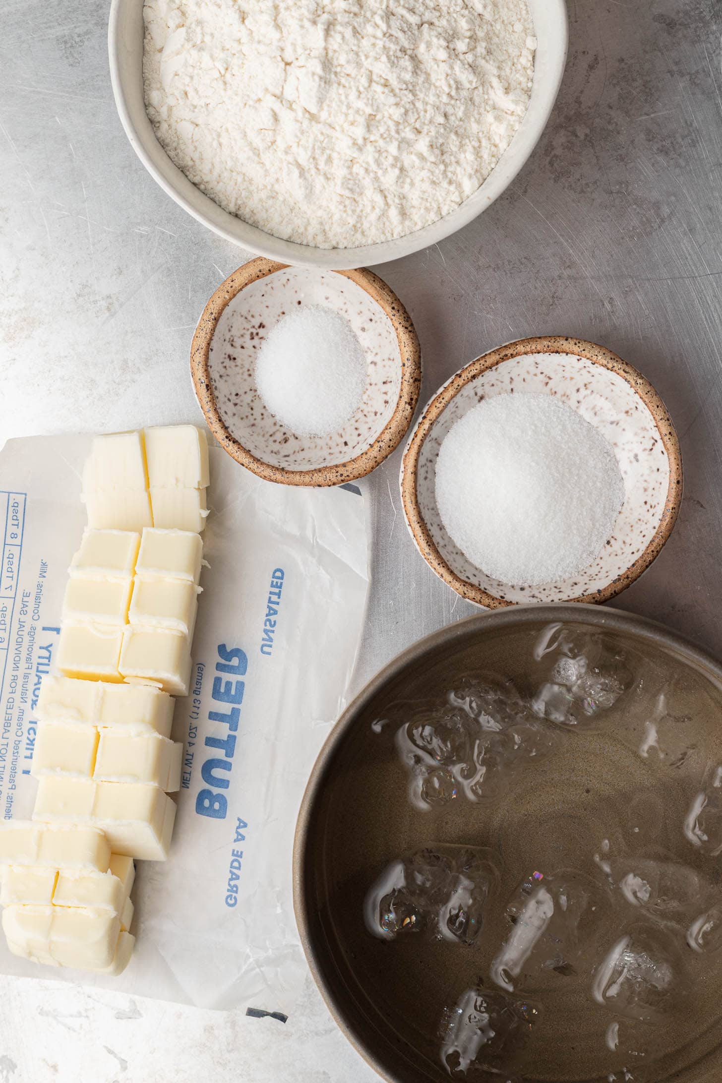 Bowls of flour, sugar, salt, cubed butter, and a bowl of ice water arranged on a light surface.