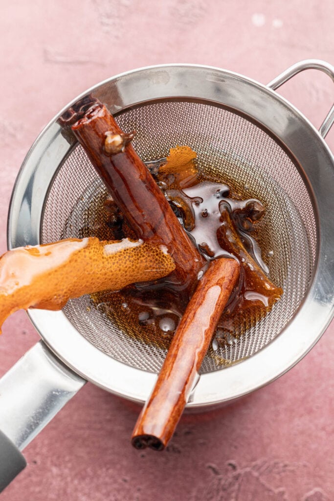Cinnamon sticks and spiced liquid being strained through a metal sieve over a pot on a pink surface.
