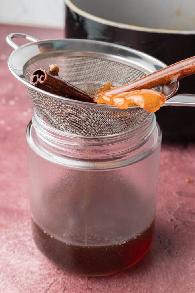 Straining spiced syrup with a cinnamon stick through a mesh sieve into a glass jar on a red surface.