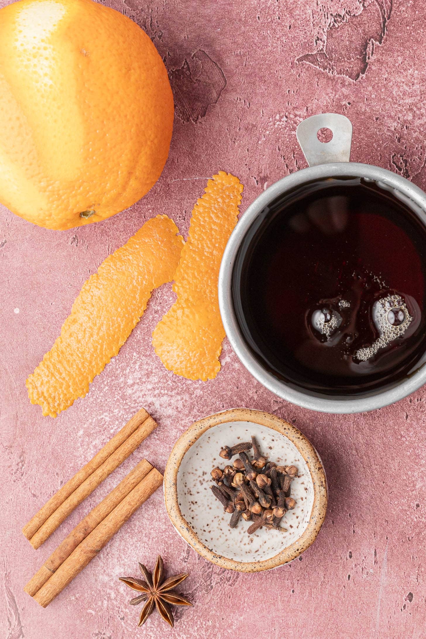 Orange, orange peel, cup of dark liquid, cinnamon sticks, cloves, and star anise on a pink surface.