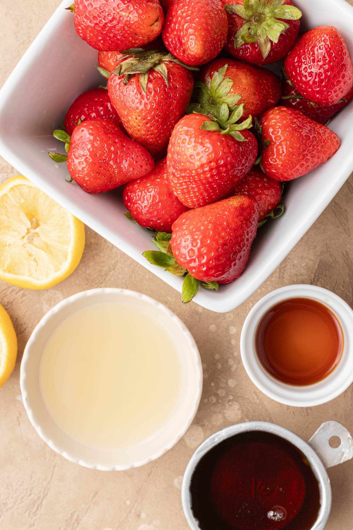 Fresh strawberries in a white bowl, with lemon halves, lemon juice, maple syrup, and vanilla in small bowls nearby.