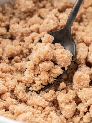 A spoon scooping crumbly brown sugar and flour streusel topping from a bowl.